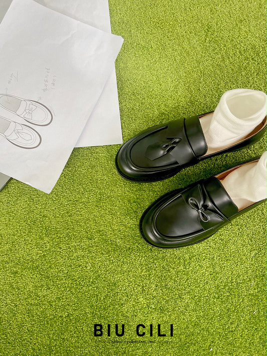 Black leather loafers for women featuring asymmetric tie and bow details, crafted in matte calfskin with low heel and soft structure, place on grass.