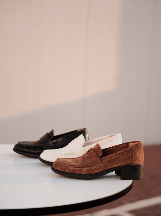 Women's loafer heels with asymmetric detail on a round white table, photographed in warm natural light, one shoe for each color, black, white and brown