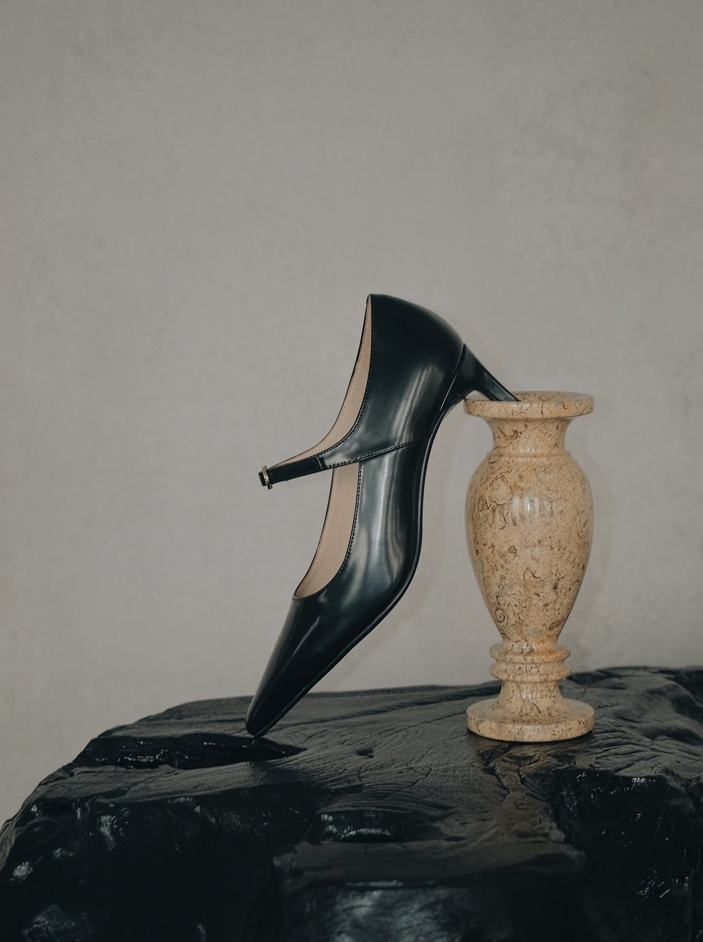 Black pointed-toe Mary Jane heel with metal buckle displayed sideway against an abstract white sculpture on a stone pedestal