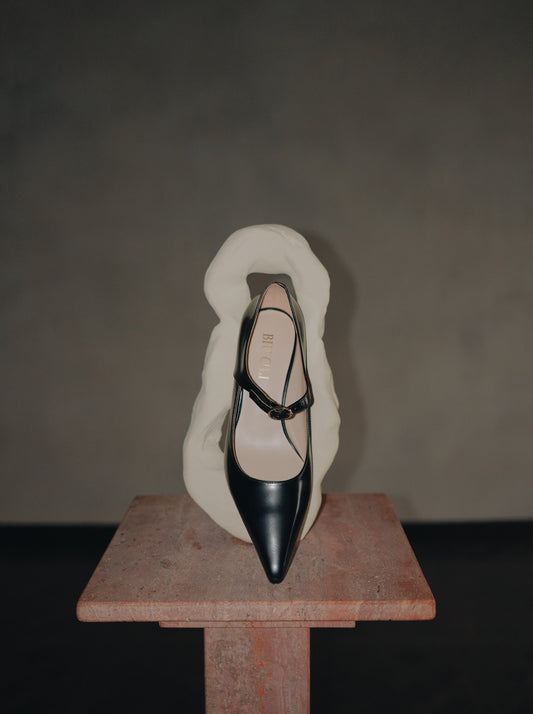 Black pointed-toe Mary Jane heel with metal buckle displayed upright against an abstract white sculpture on a stone pedestal