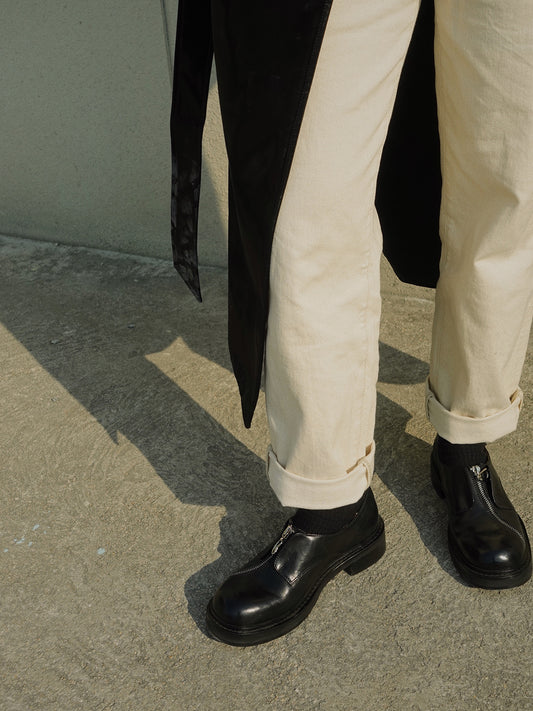 Model wearing black asymmetric leather loafers with zipper detail, paired with cream cuffed pants and a black coat, photographed outdoors on concrete.
