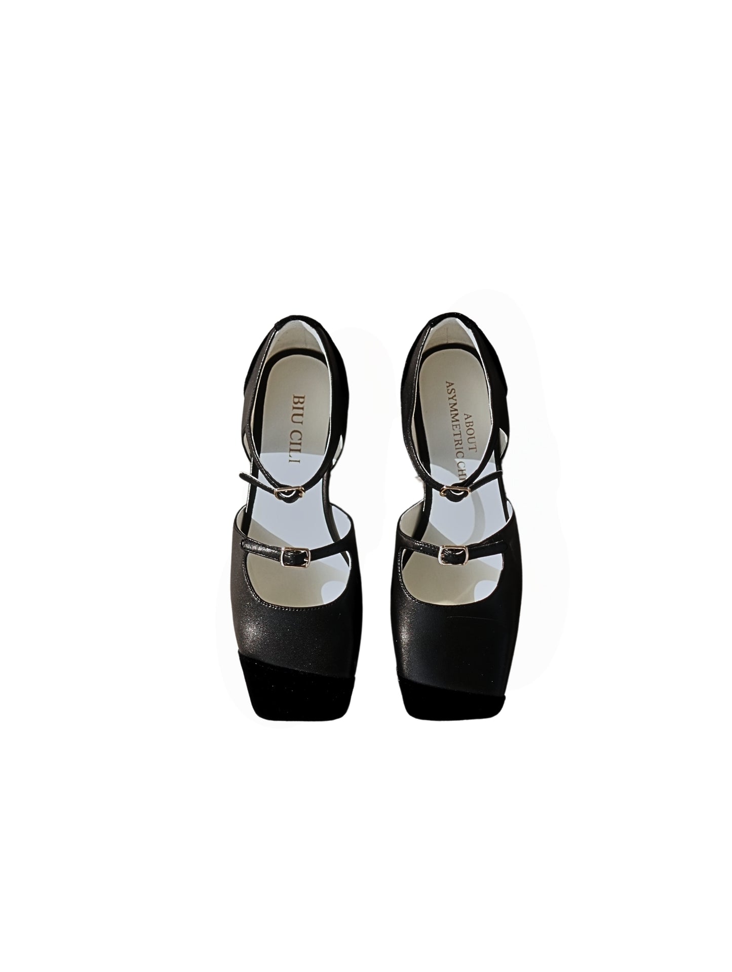 A pair of black leather mary jane flats with straps and asymmetric velvet details at toe front, placed on a white background