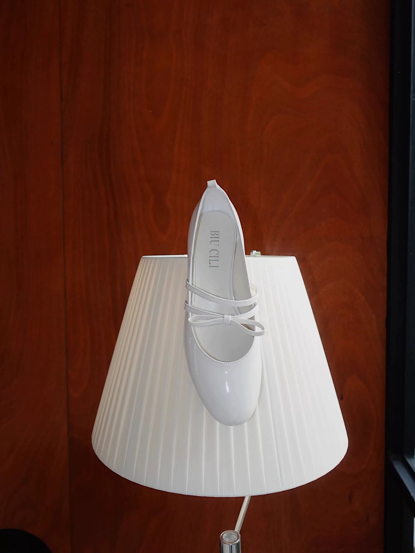 White patent leather Mary Jane heels with asymmetric bow straps, placed on a lamp