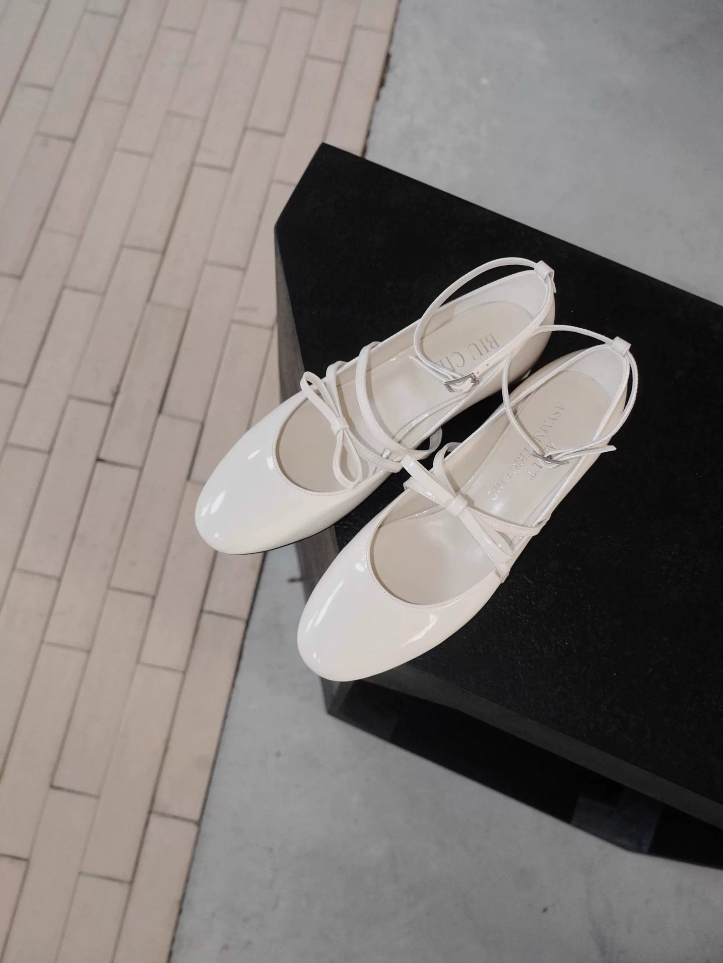 White patent leather Mary Jane heels with asymmetric bow straps, displayed on a dark upholstered chair in natural light
