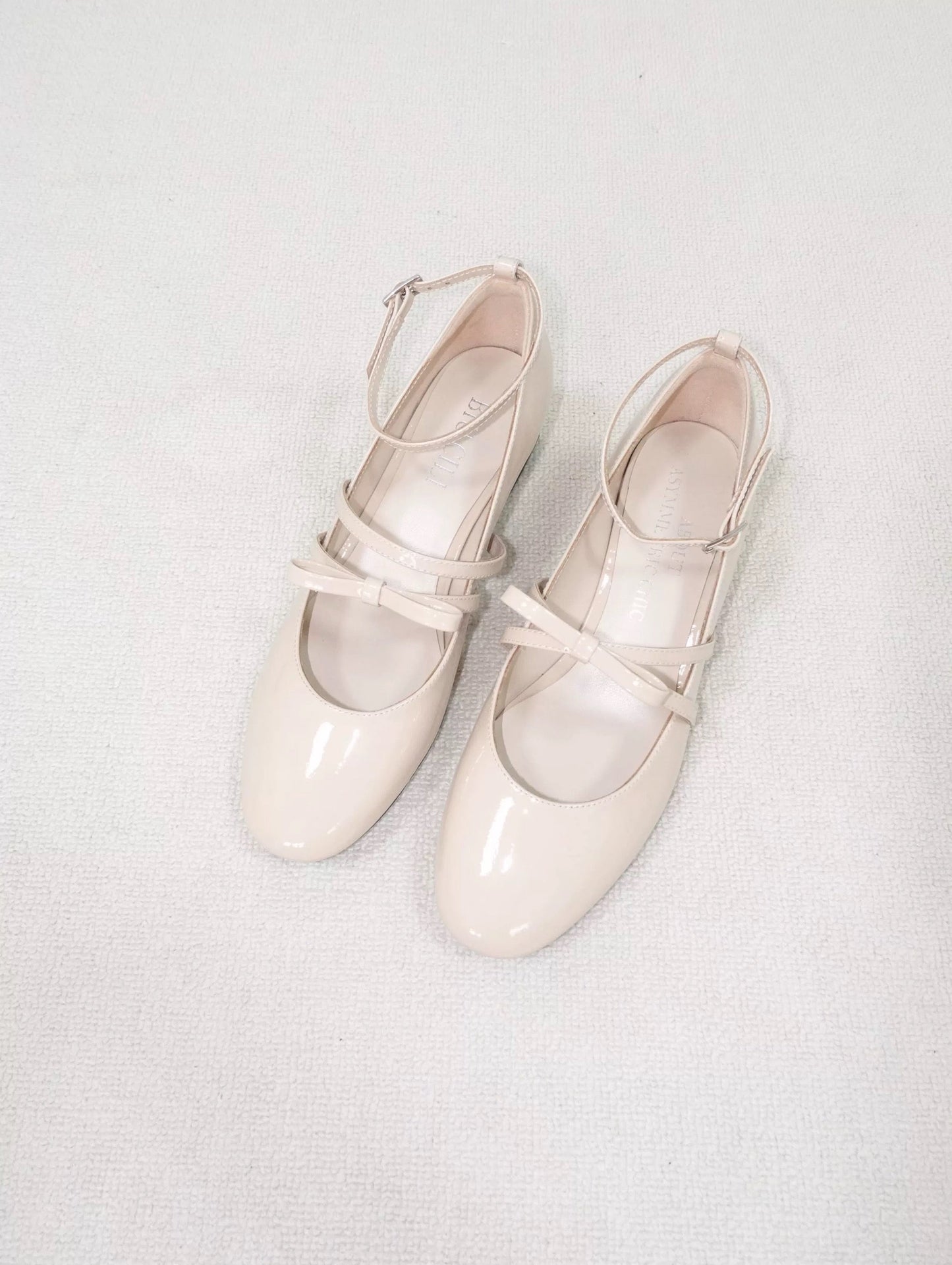 Beige patent leather Mary Jane heels with asymmetric bow straps, placed on the carpet floor