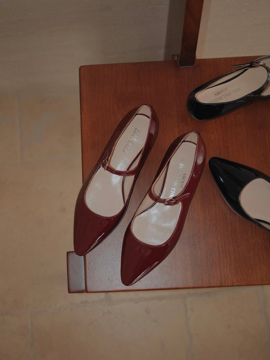 Red patent leather pointed-toe Mary Jane flats on a wooden surface, with a blurred black pair in the background