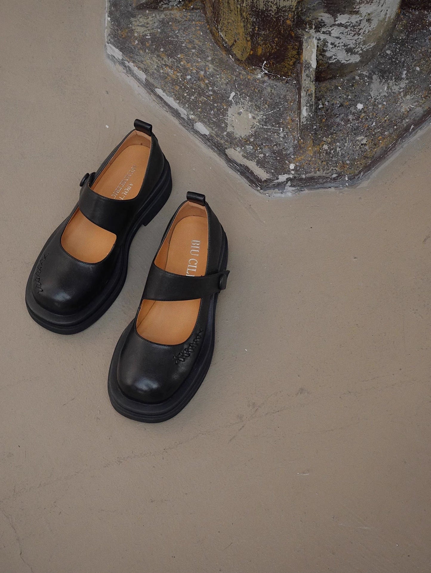 Black platform Mary Jane shoes with wide strap and low block heel, displayed on a textured concrete floor beside a wall