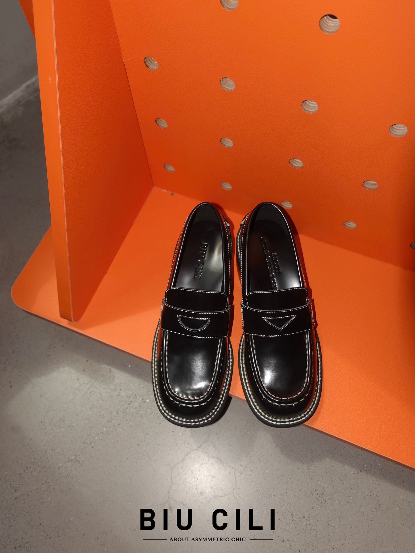A pair of black leather loafer heels with asymmetric details for women, placed on a highly contrasted orange surface.