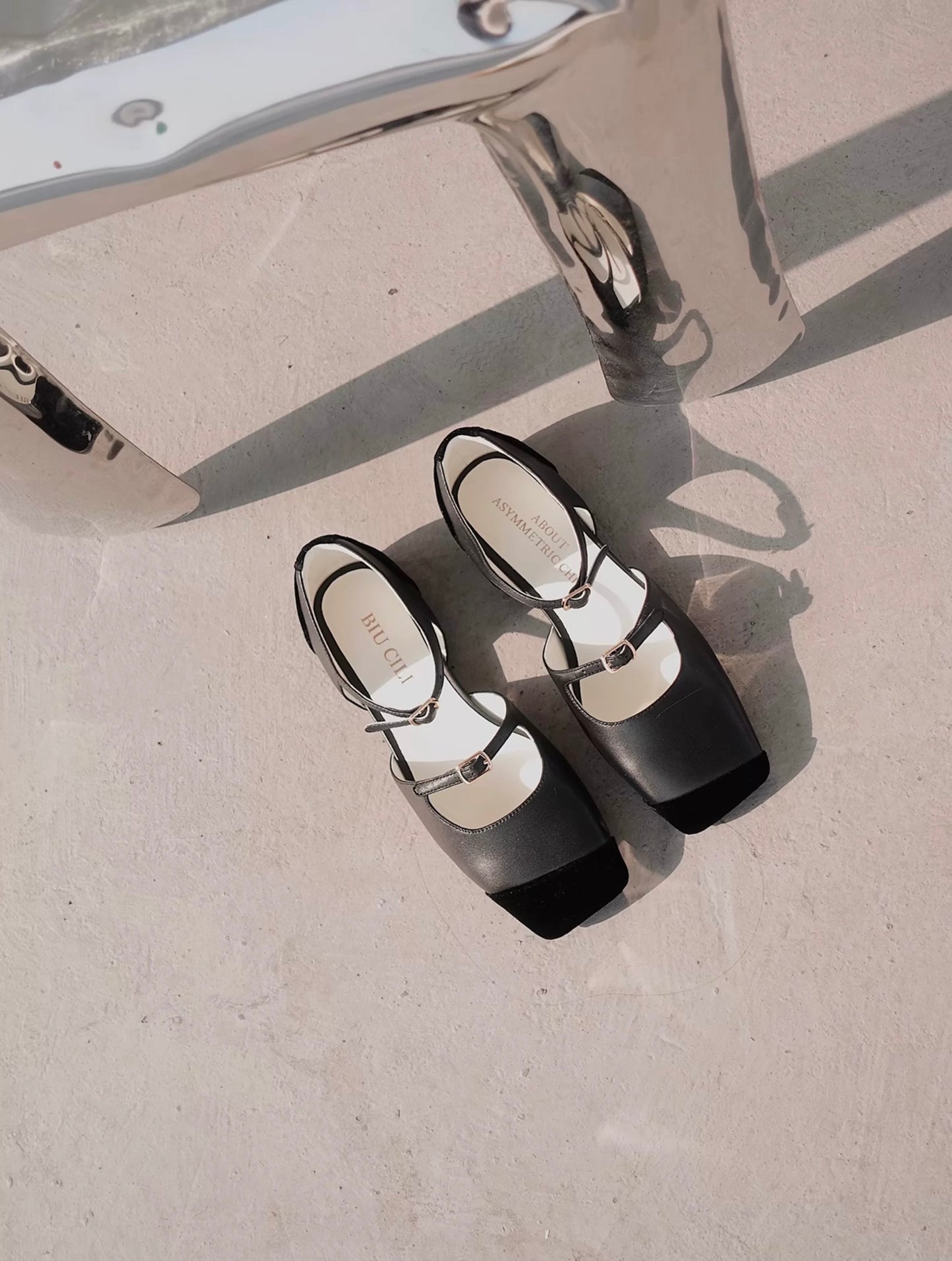 Top view of a pair of black leather mary jane flats with straps and asymmetric velvet details at toe front, placed on the floor next to a chair
