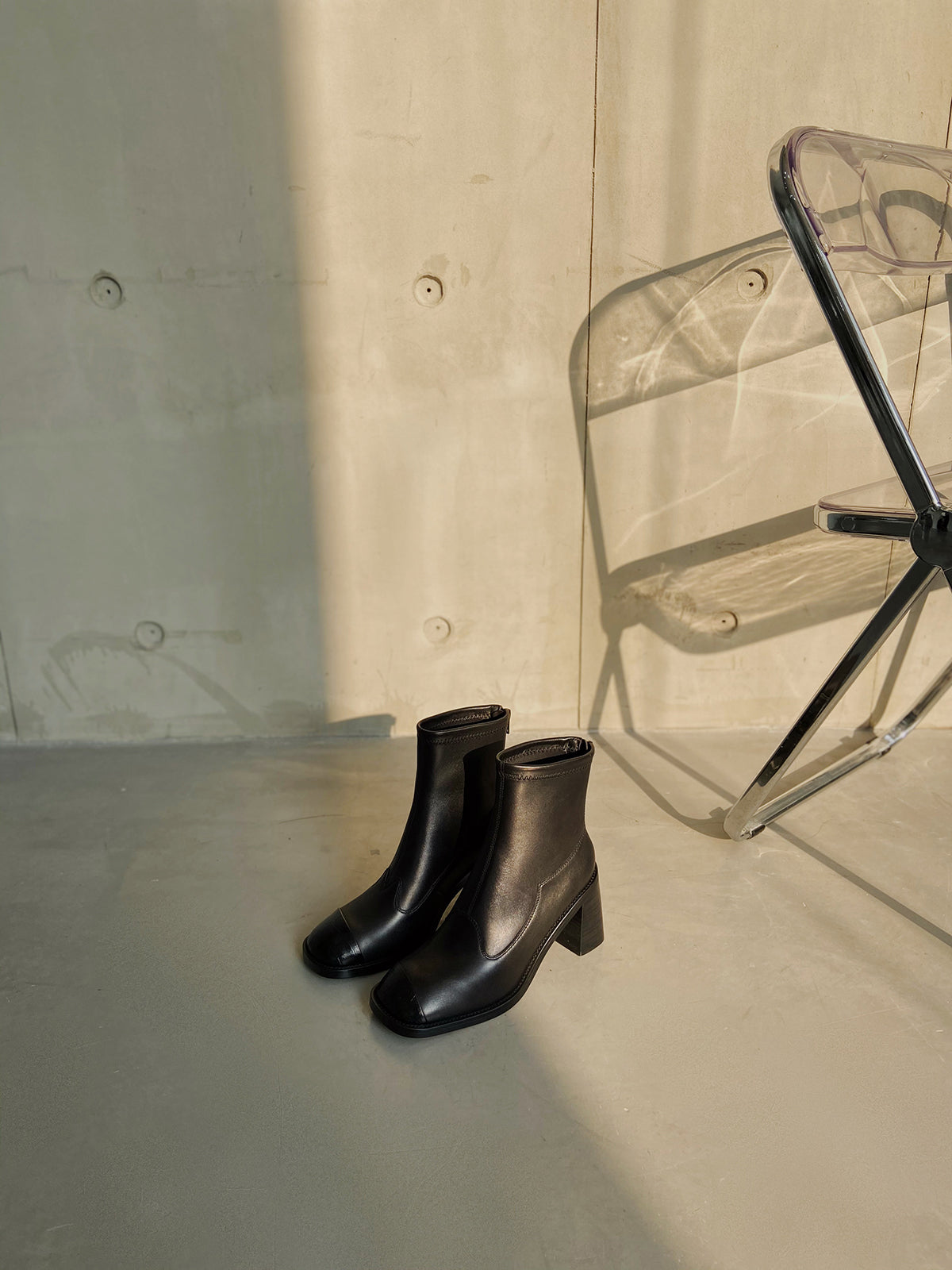 Women's black leather square-toe ankle boots with block heels, styled against a concrete wall in warm natural light