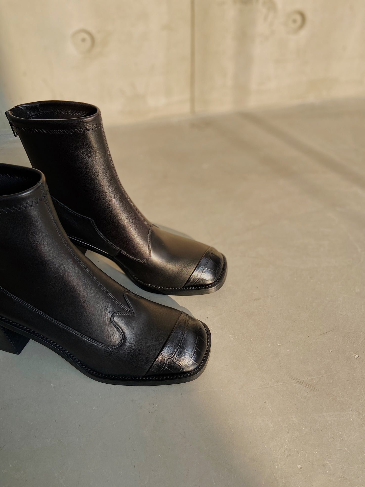 Women's black leather square-toe ankle boots with block heels, styled against a concrete wall in warm natural light