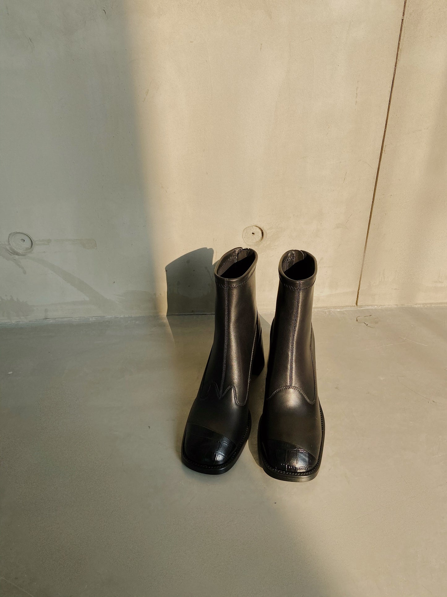 Women's black leather square-toe ankle boots with block heels, styled against a concrete wall in warm natural light