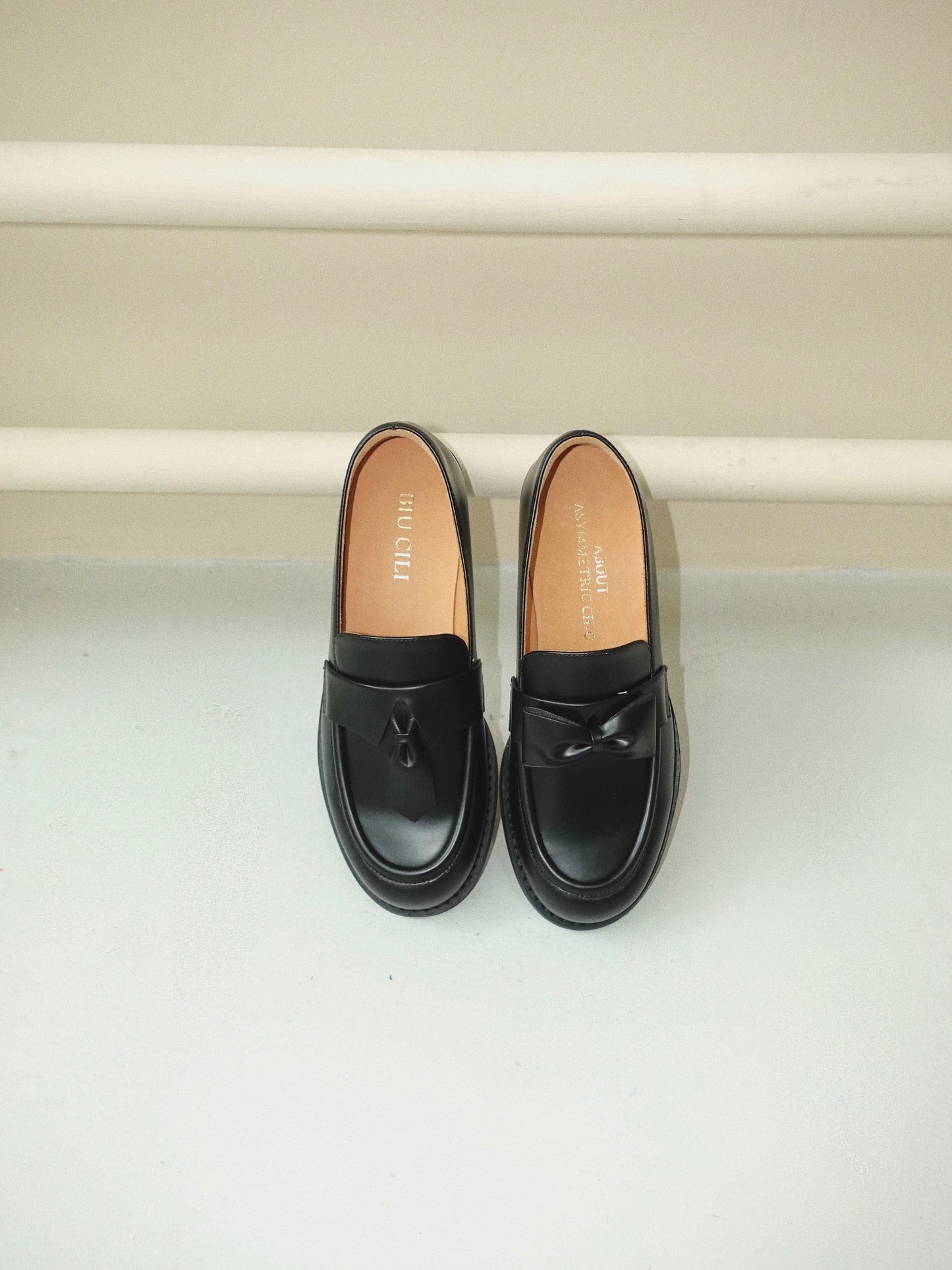 Black leather loafers for women featuring asymmetric tie and bow details, crafted in matte calfskin with low heel and soft structure, placed on the ground.