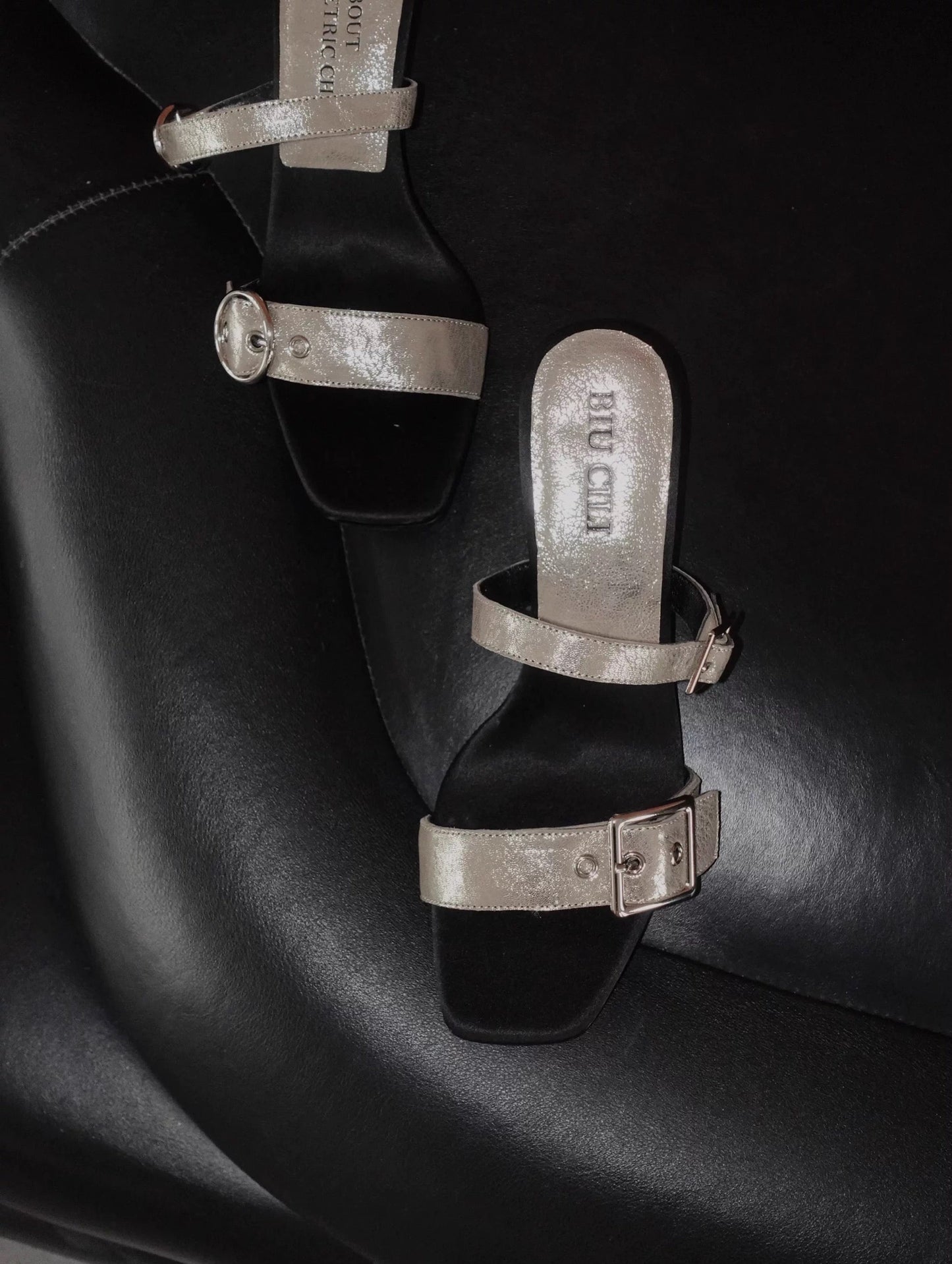 A mid heel sandal with two thick leather straps and shiny silver buckle - top view - placing on a black leather surface