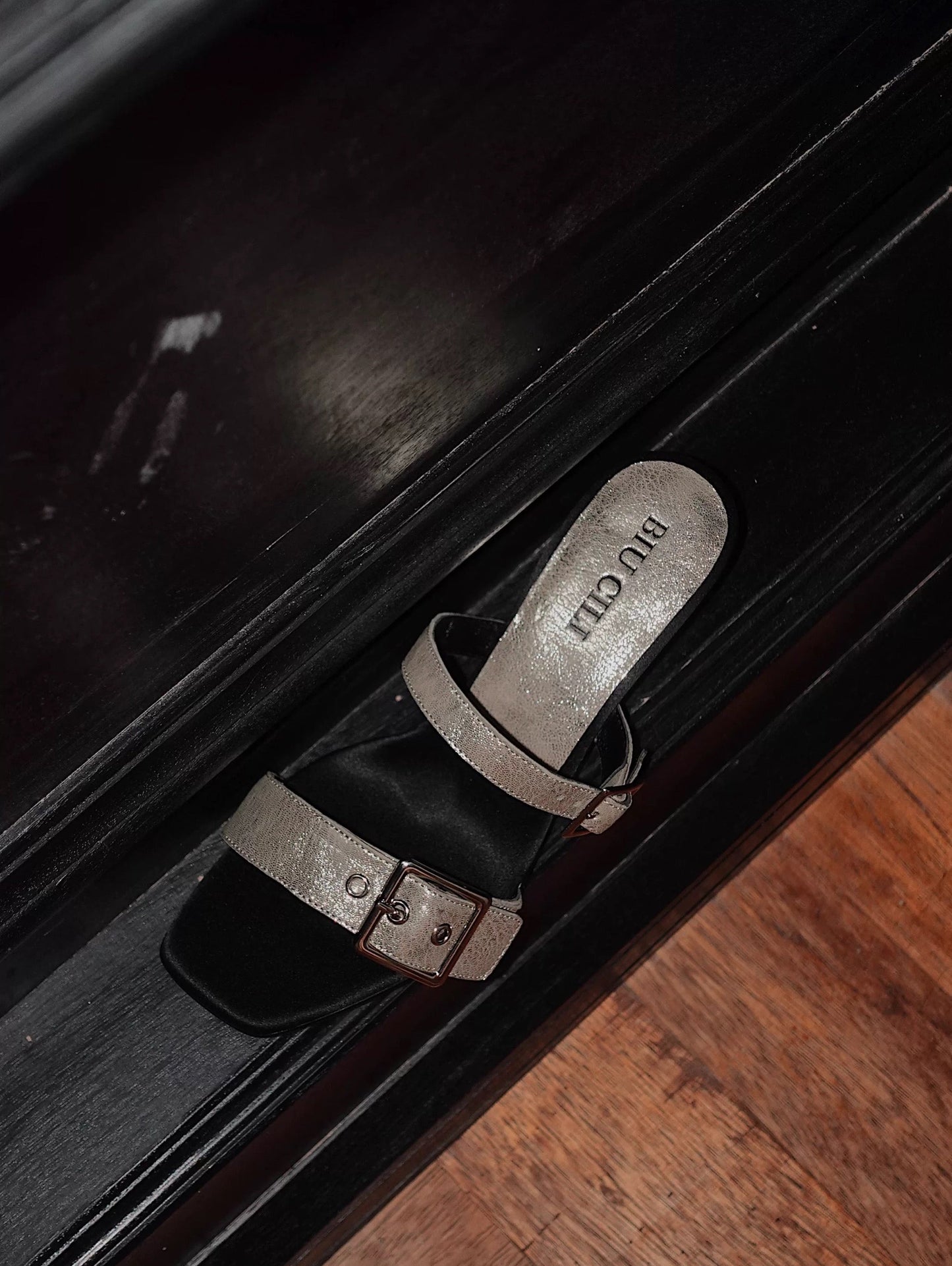 A mid heel sandal with two thick leather straps and shiny silver buckle - side view - placing on a black leather surface