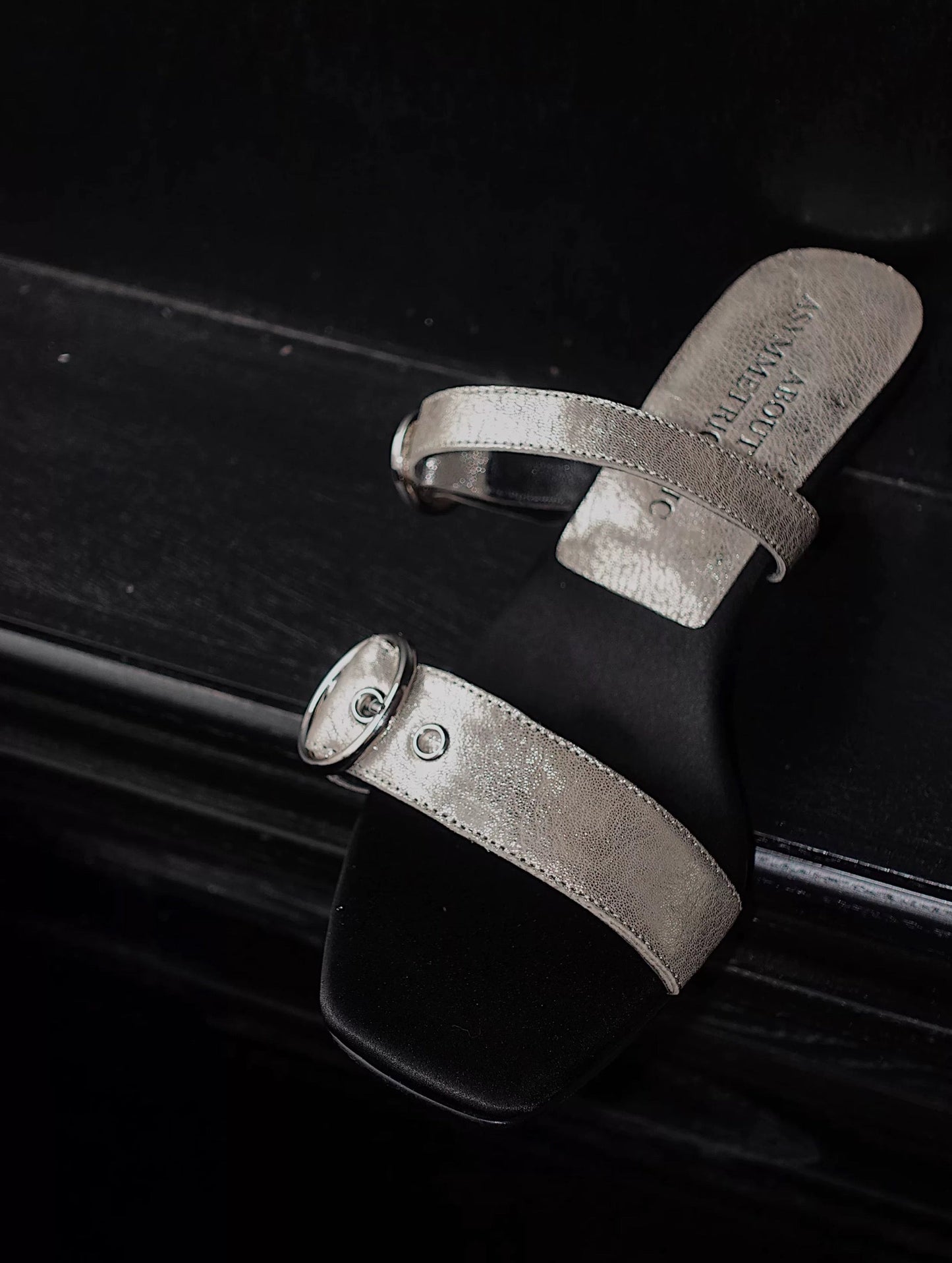 A close view of a mid heel sandal with two thick leather straps and shiny silver buckle - top view - placing on a black leather surface