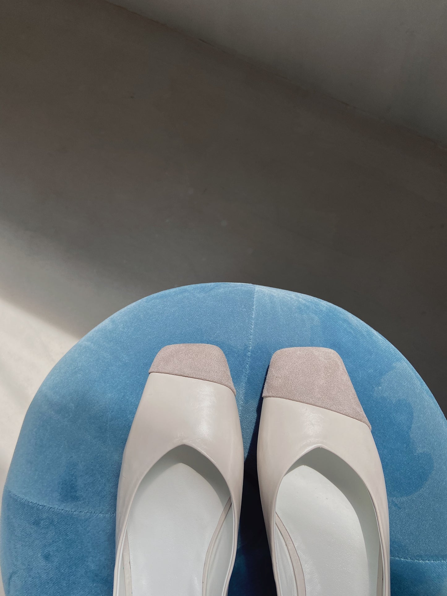 Front view of square-toe Mary Jane heels in white leather with light grey suede toe caps, resting on a blue upholstered surface