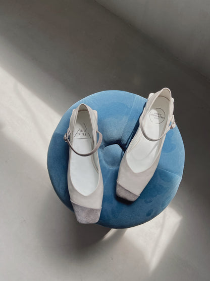 Top view of white leather Mary Jane heels with grey suede ankle straps, placed on a soft blue circular stool