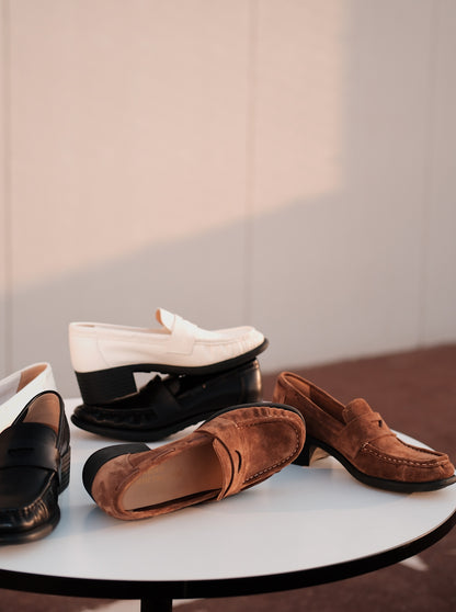 Four women's mid-heel loafers with asymmetric detail in brown and white,on a round white table, photographed in warm natural light
