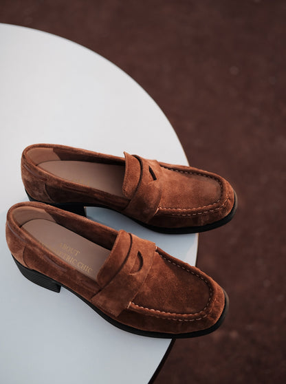 Women's brown suede mid-heel loafers with asymmetric detail on a round white table, photographed in warm natural light