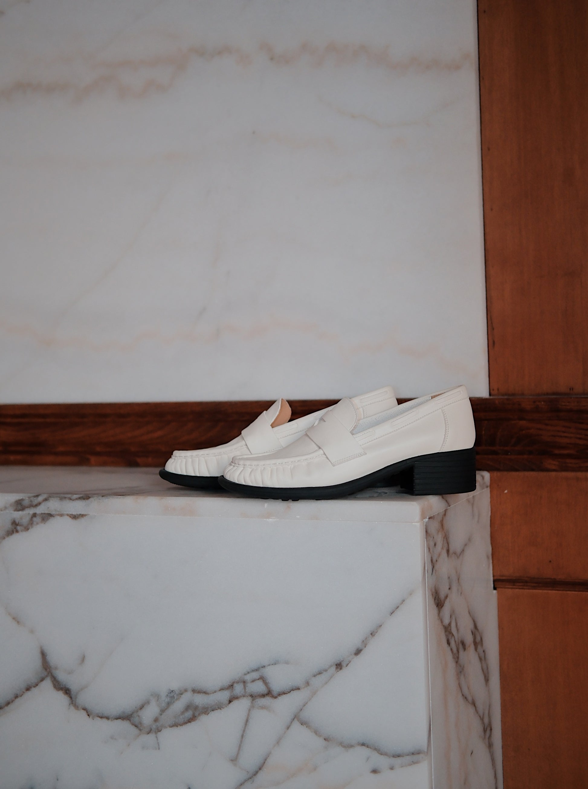 Women's white leather loafer heels with asymmetric detail, placed on the floor