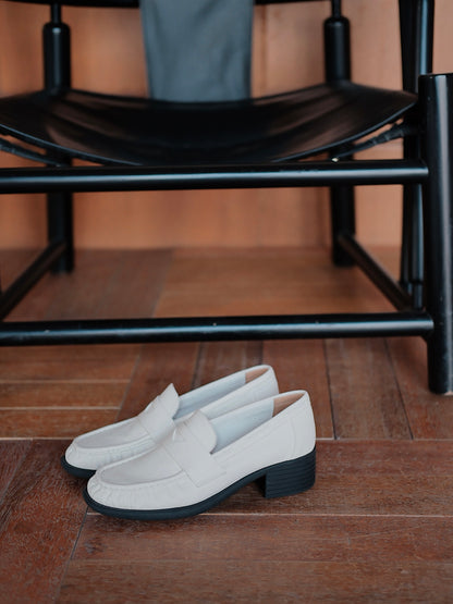 Women's white leather loafer heels with asymmetric detail, placed on the floor