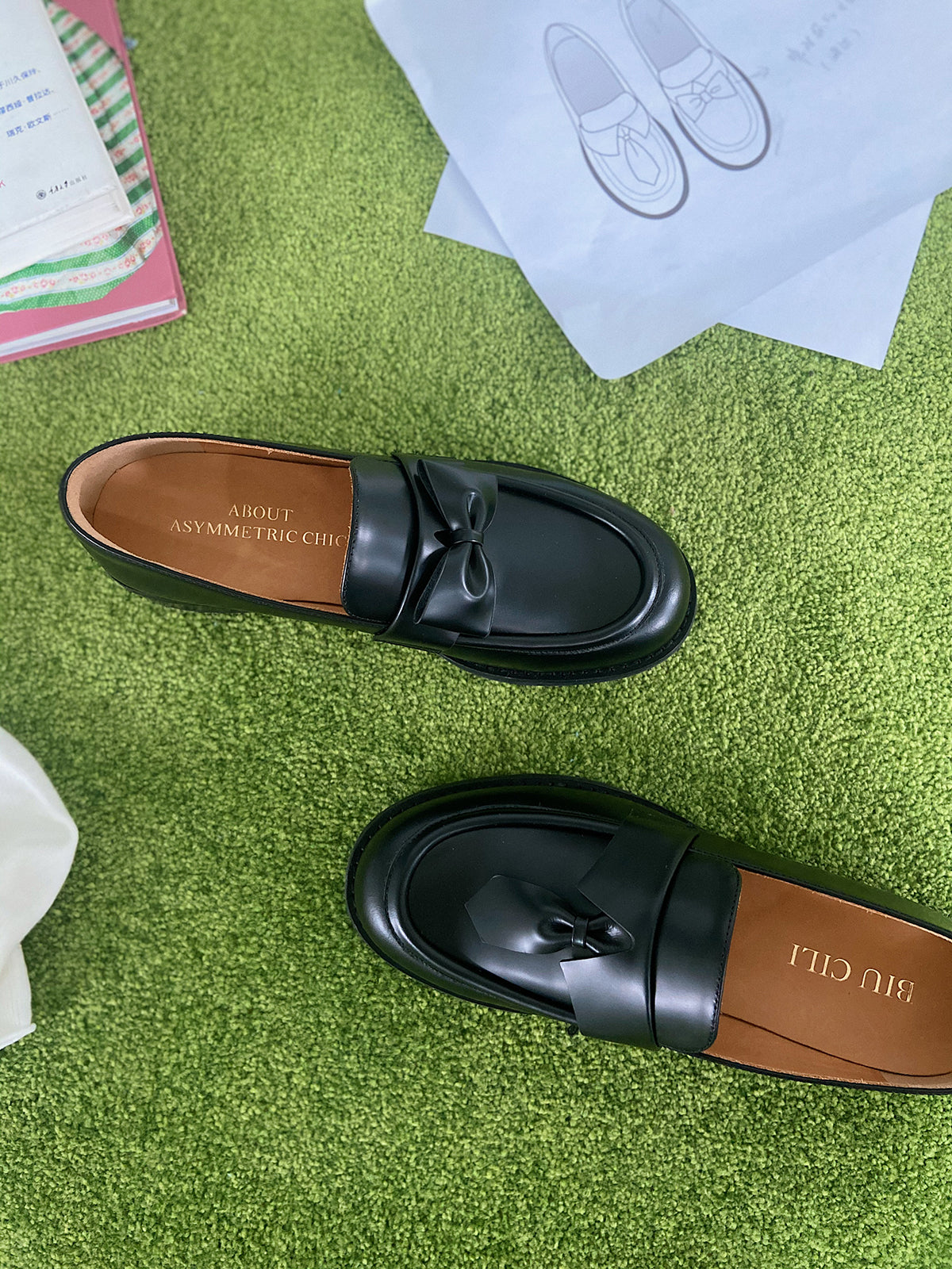 Black leather loafers for women featuring asymmetric tie and bow details, crafted in matte calfskin with low heel and soft structure, placed on grass.