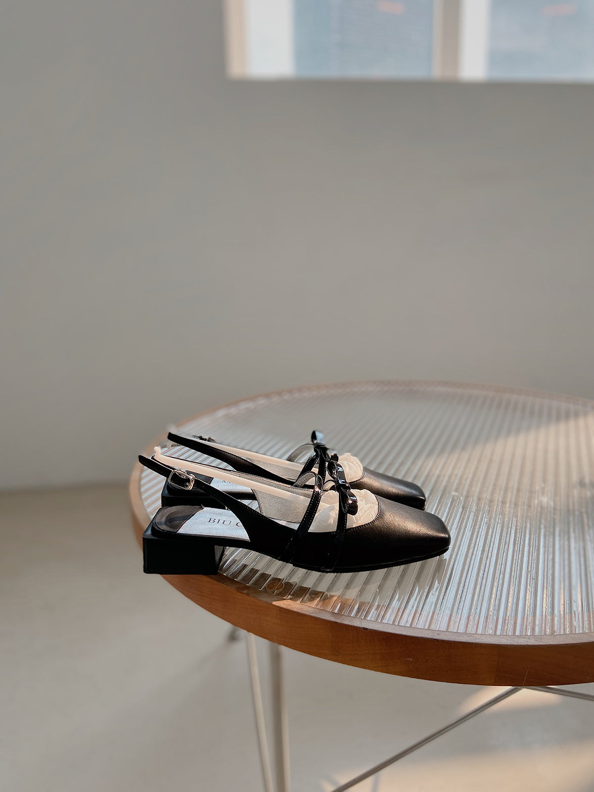 Black square toe Mary Jane shoes with asymmetric bow and low block heel, photographed on a glass table