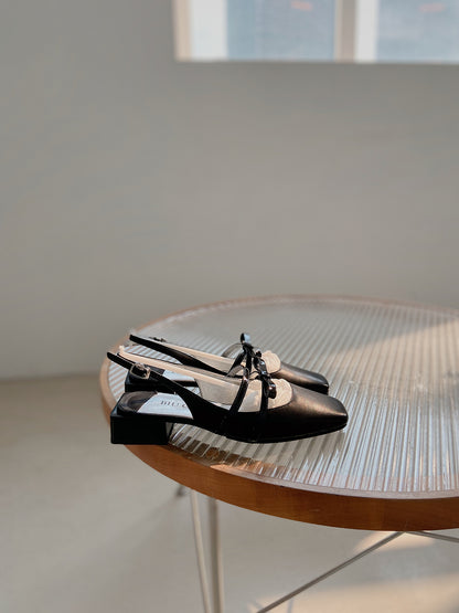 Black square toe Mary Jane shoes with asymmetric bow and low block heel, photographed on a glass table