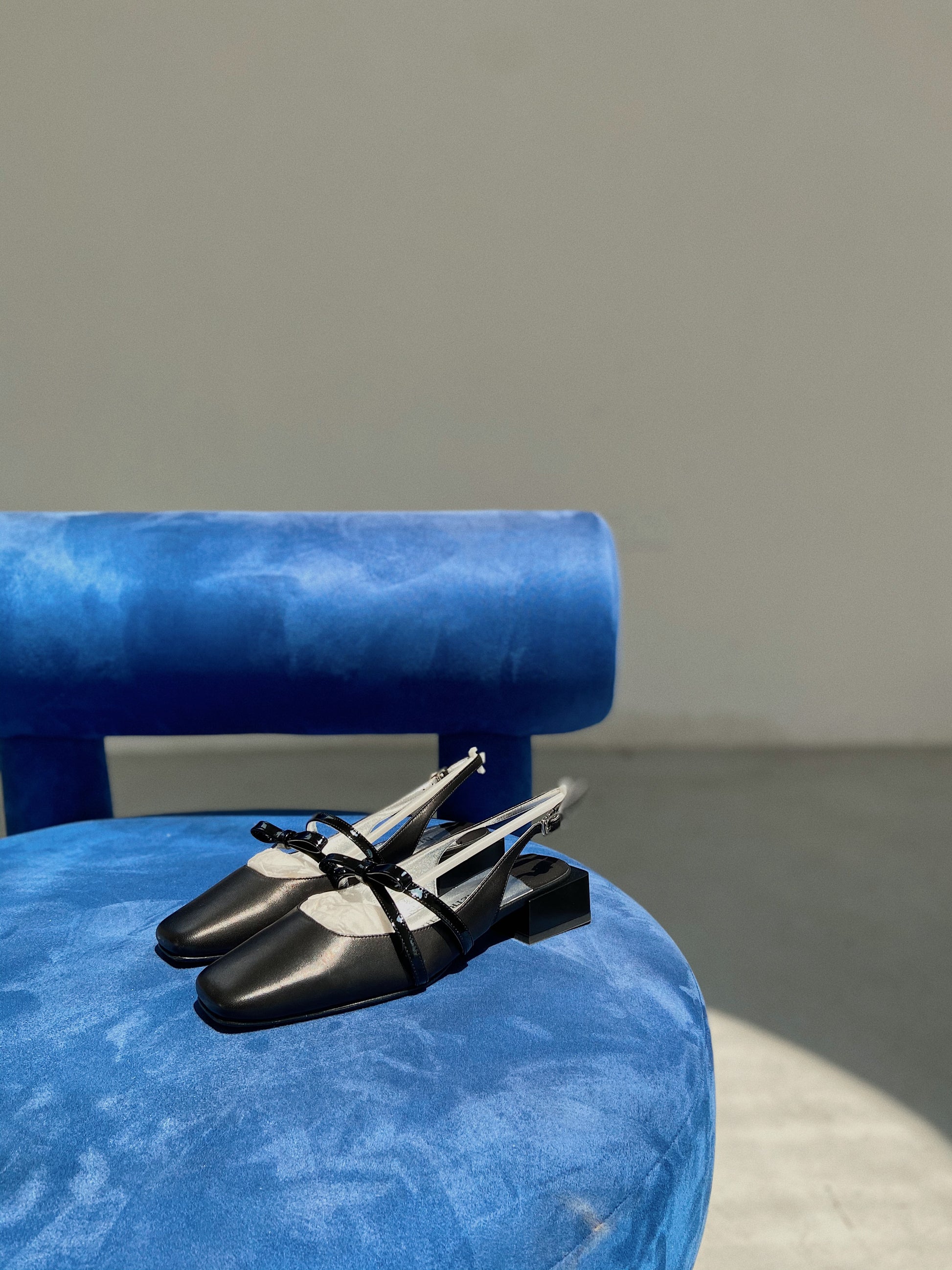 Black square toe Mary Jane shoes with asymmetric bow and low block heel, photographed on a blue chair