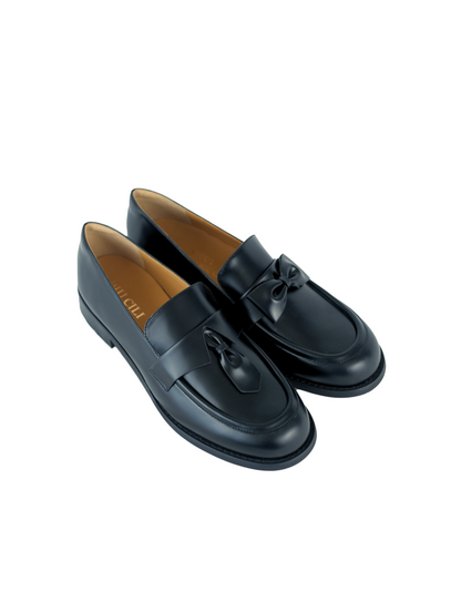 Black leather loafers for women featuring asymmetric tie and bow details, crafted in matte calfskin with low heel and soft structure, shown on white background.