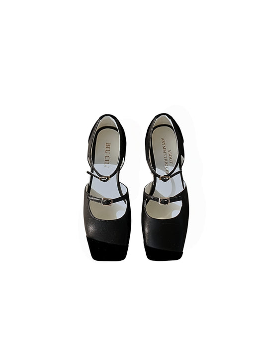 A pair of black leather mary jane flats with straps and asymmetric velvet details at toe front, placed on a white background