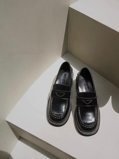 A pair of black leather loafer heels with asymmetric details for women, placing on the floor.