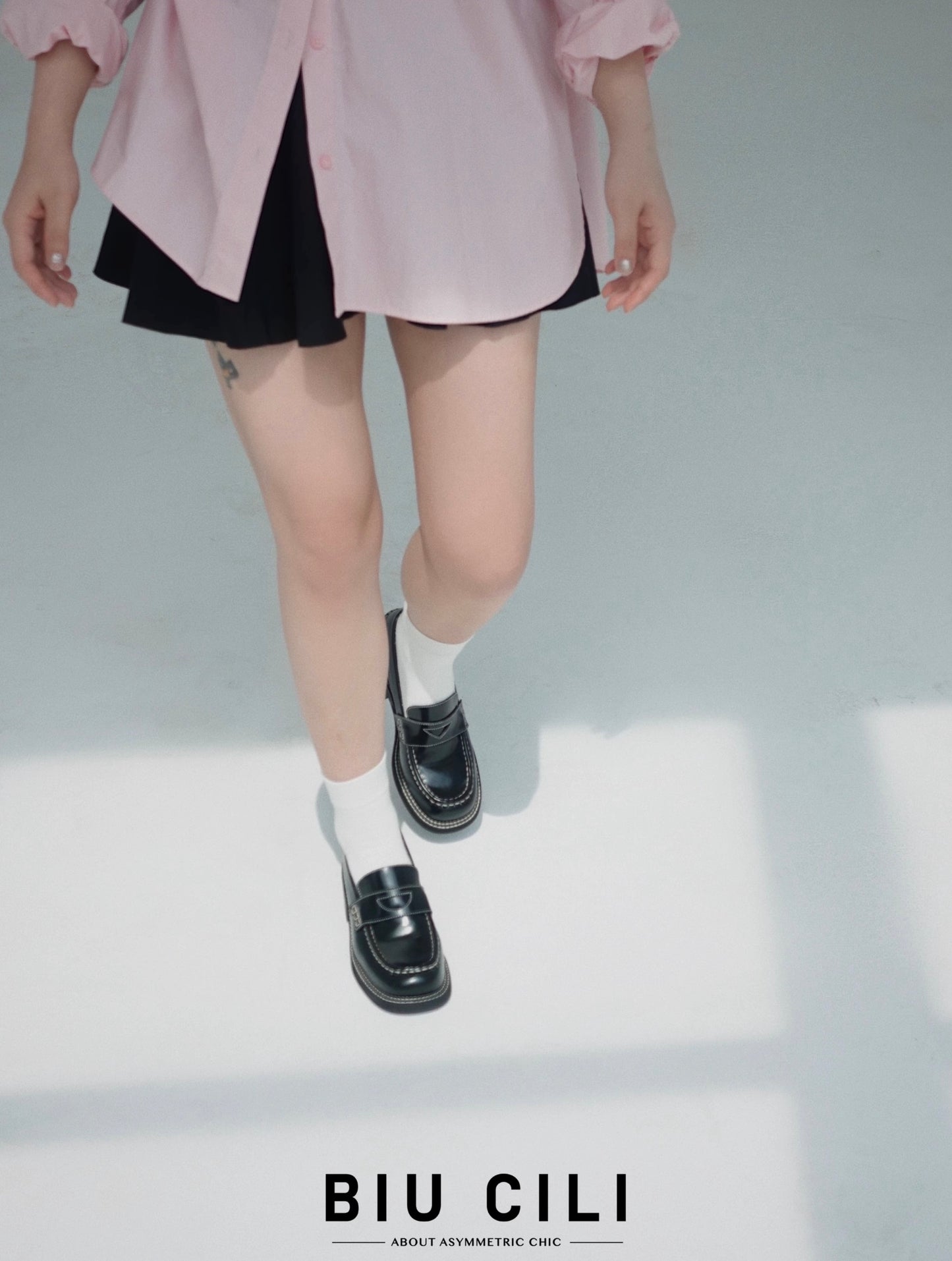 Woman wearing a pair of black leather loafer heels with white socks underneath, pairing with pink shirt top and black shorts, walking confidently