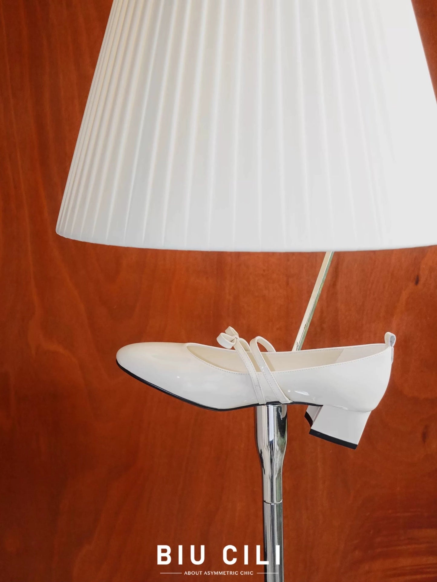 White patent leather Mary Jane heels with asymmetric bow straps, placed near a lamp