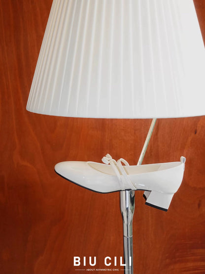 White patent leather Mary Jane heels with asymmetric bow straps, placed near a lamp