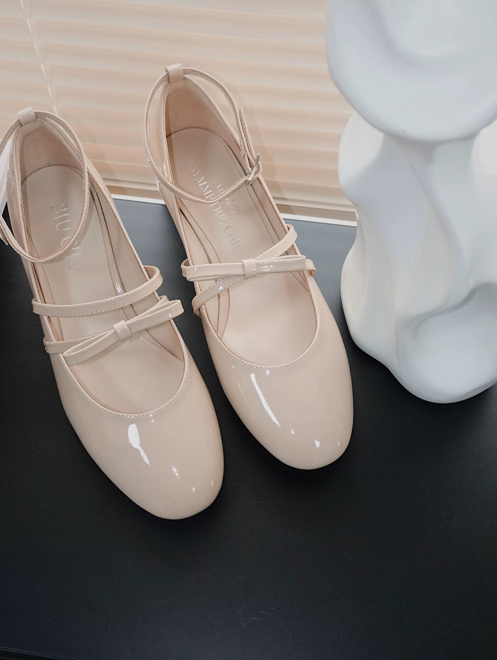 Beige patent leather Mary Jane heels with asymmetric bow straps, placed beside a white abstract sculpture on a dark tabletop