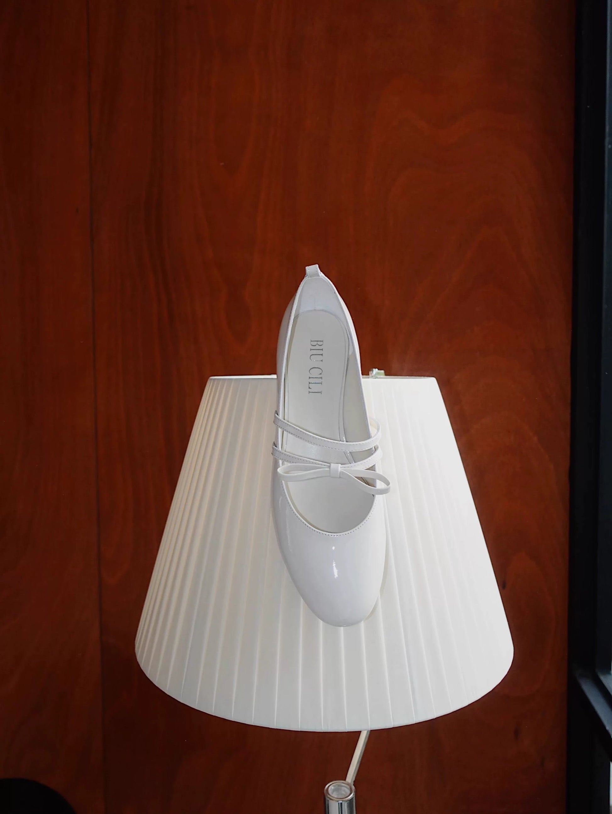 White patent leather Mary Jane heels with asymmetric bow straps, placed on a lamp