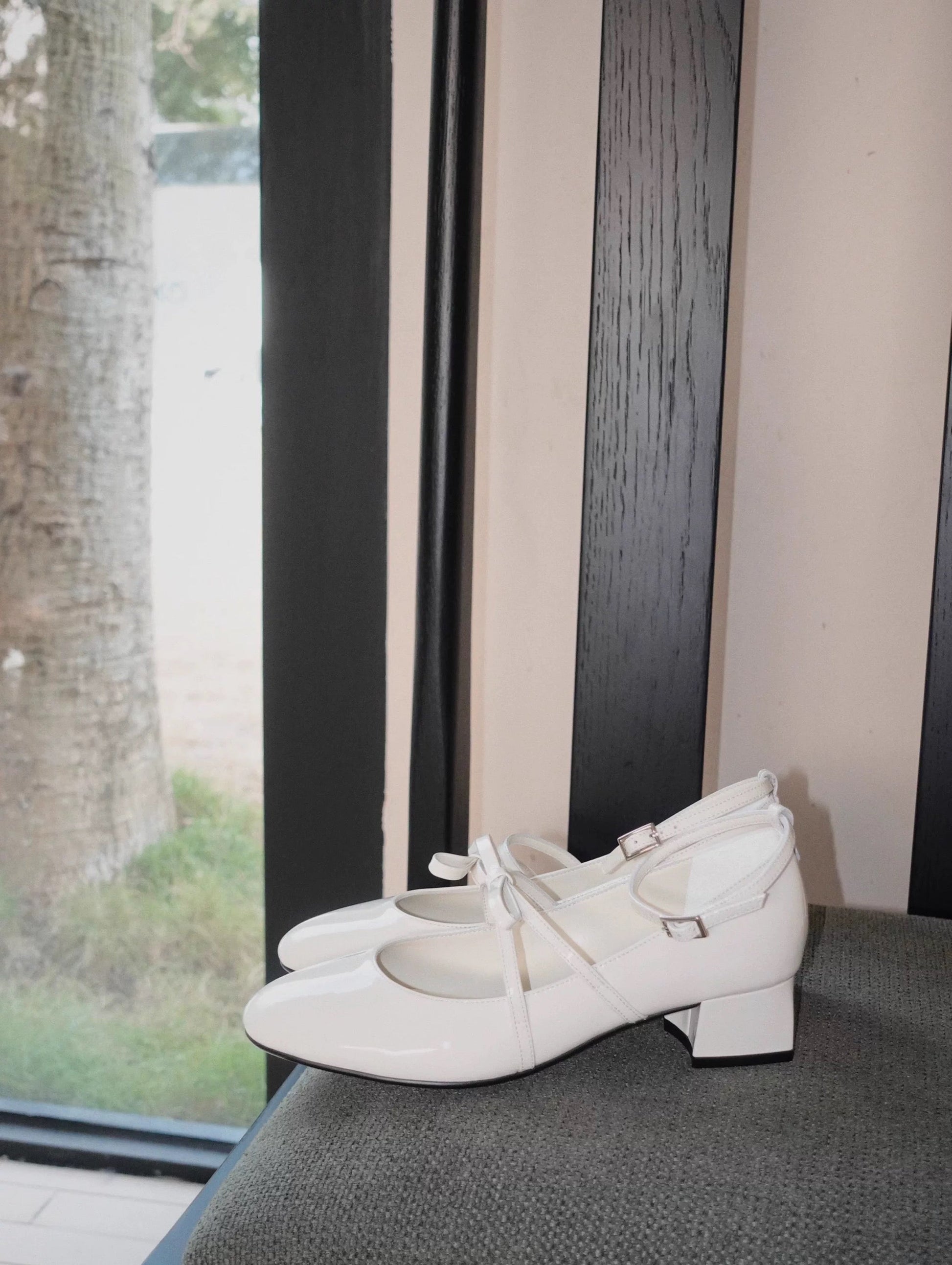 White patent leather Mary Jane heels with asymmetric bow straps, displayed on a dark chair in natural light