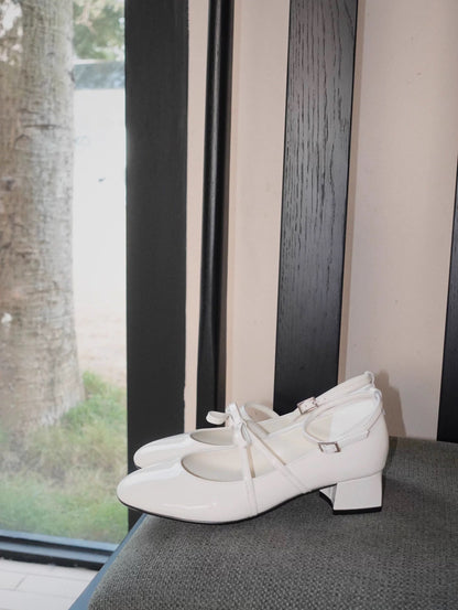 White patent leather Mary Jane heels with asymmetric bow straps, displayed on a dark chair in natural light