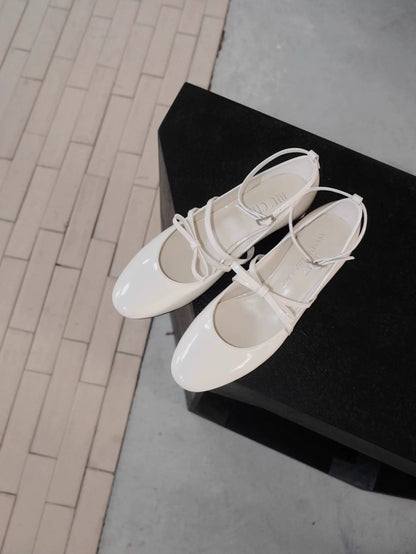 White patent leather Mary Jane heels with asymmetric bow straps, displayed on a dark upholstered chair in natural light