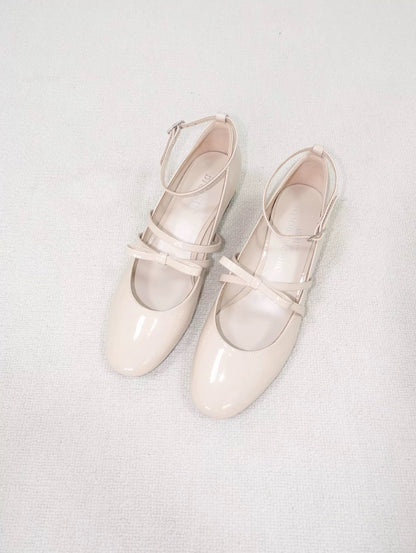 Beige patent leather Mary Jane heels with asymmetric bow straps, placed on the carpet floor