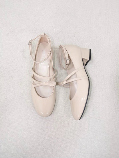 Beige patent leather Mary Jane heels with asymmetric bow straps, placed on the carpet floor