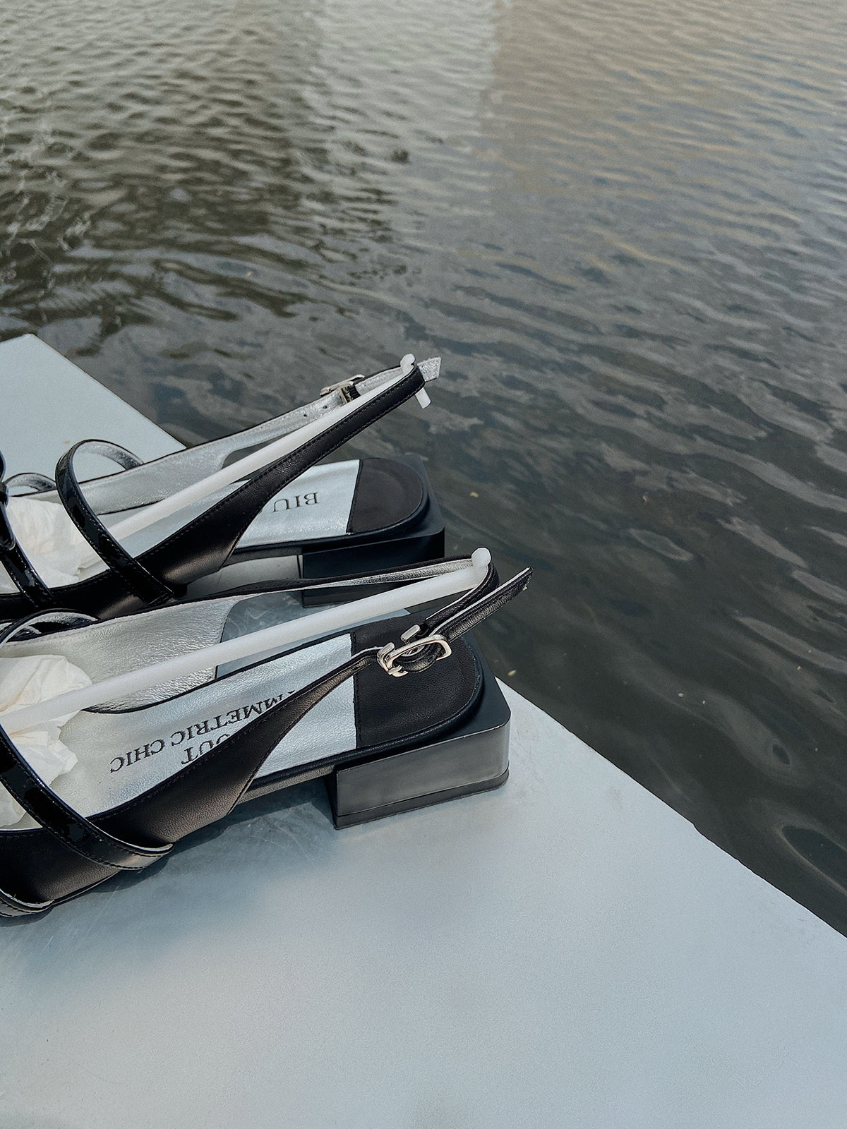 Black square toe Mary Jane shoes with asymmetric bow and low block heel, photographed near water on a white surface.