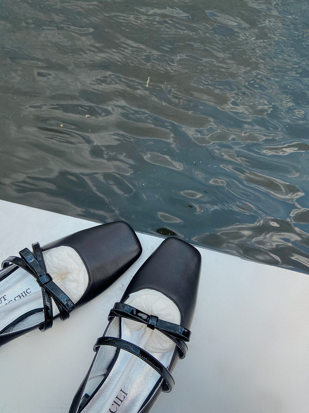 Black square toe Mary Jane shoes with asymmetric bow and low block heel, photographed near water on a white surface.