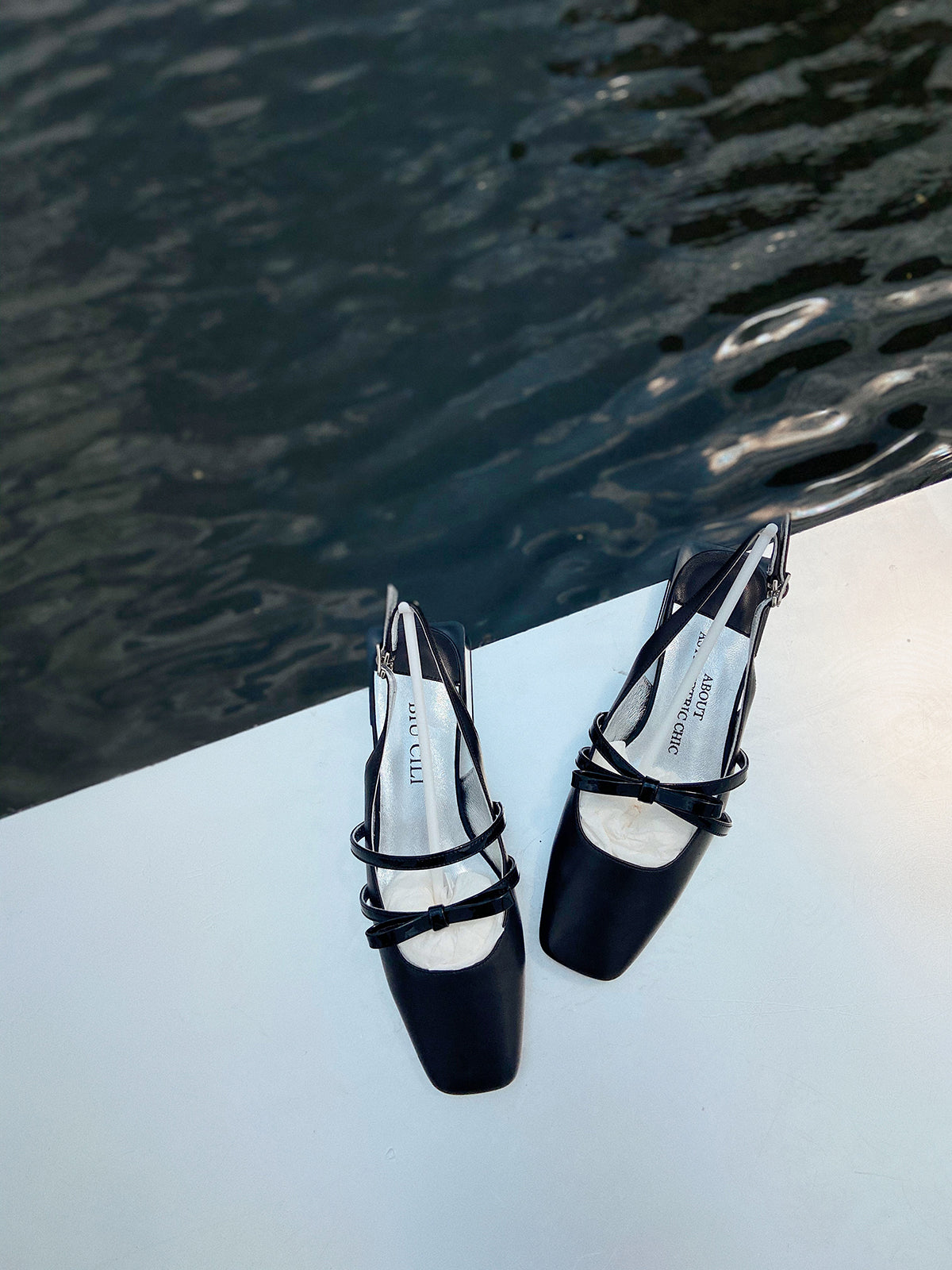Black square toe Mary Jane shoes with asymmetric straps, bow detials and low block heel, photographed near water on a white surface.