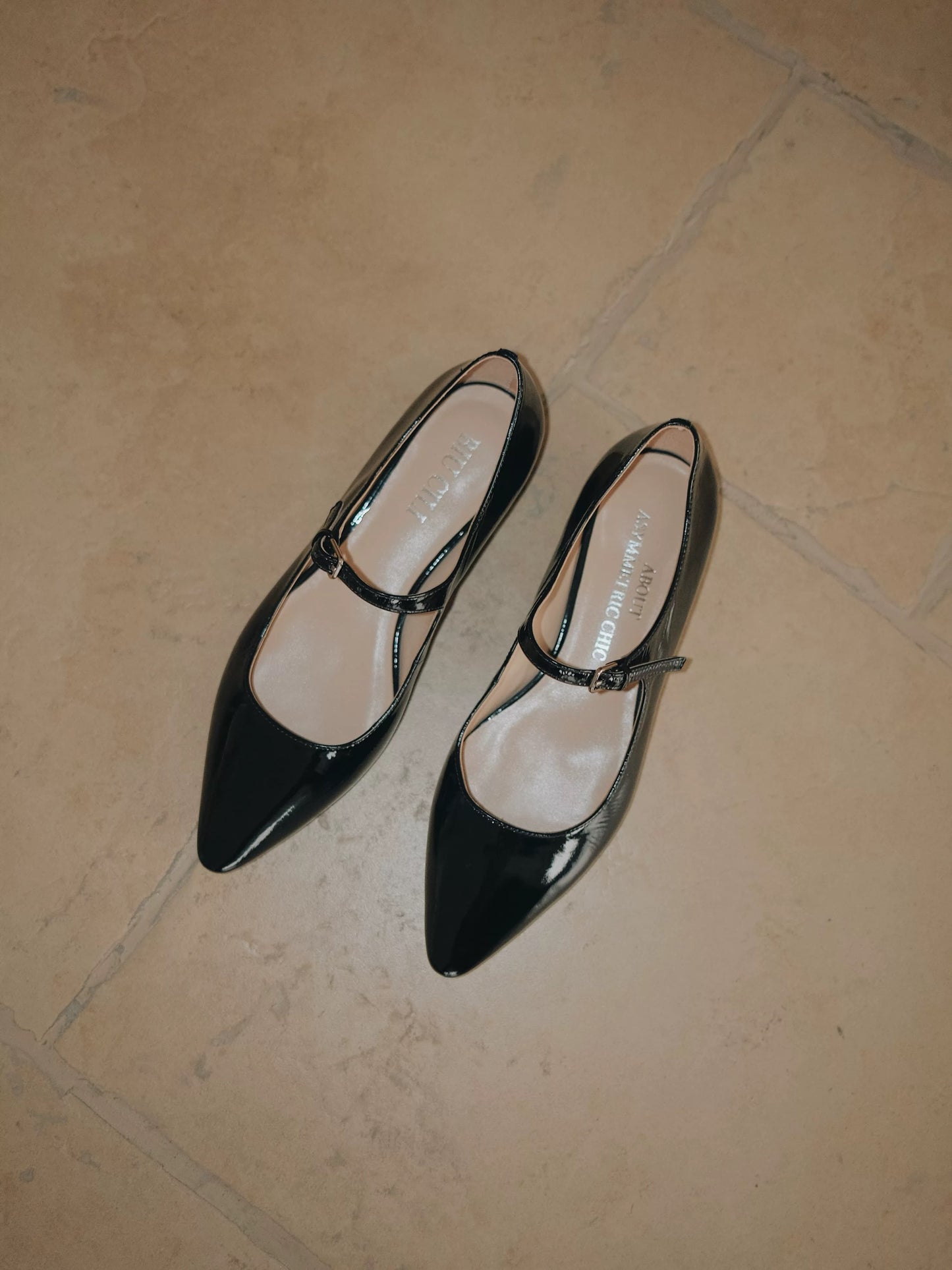 A pair of glossy balck patent leather pointed-toe Mary Jane flats with a single strap, displayed on the ground