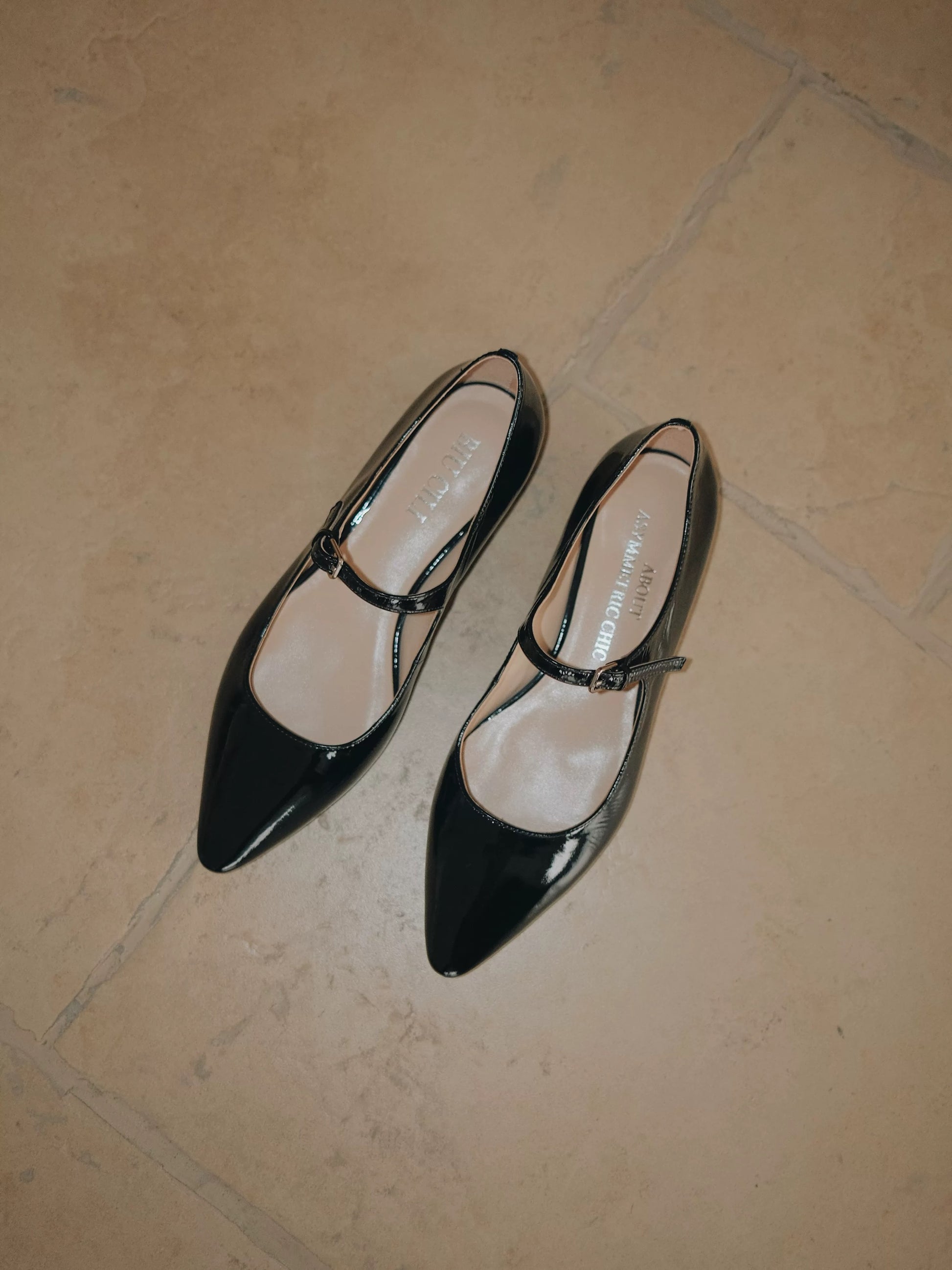 A pair of glossy balck patent leather pointed-toe Mary Jane flats with a single strap, displayed on the ground