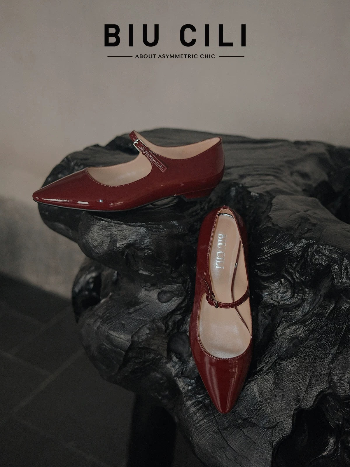 Pair of merlot red patent leather pointed-toe Mary Jane flats with a single strap, displayed on a sculptural black wooden surface