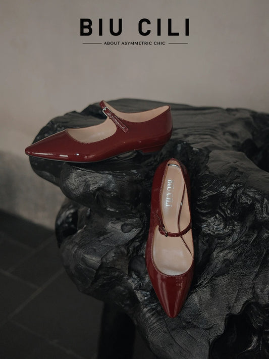 Pair of merlot red patent leather pointed-toe Mary Jane flats with a single strap, displayed on a sculptural black wooden surface